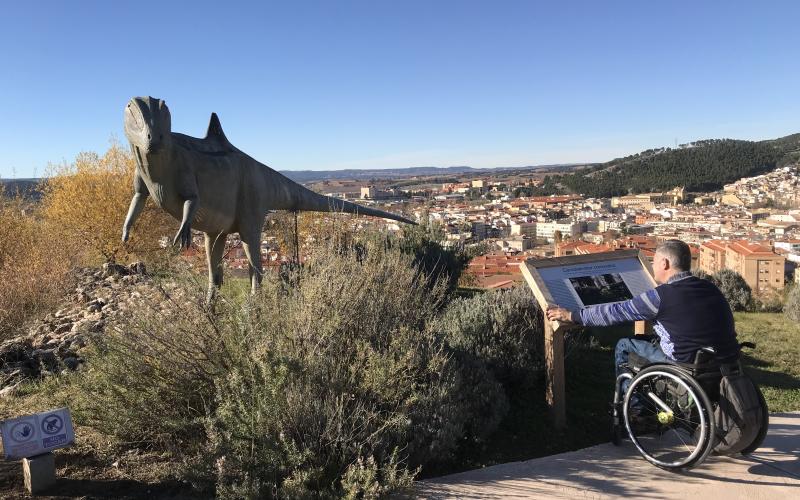 Usuario de silla de ruedas mirando una reproducción de Dinosaurio en los senderos exteriores del  MUPA